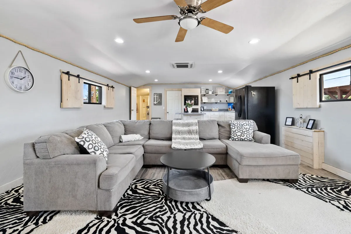 A spacious living room featuring a large sectional sofa, zebra print rug, coffee table, and a wall clock. The room is decorated with rustic touches and invites guests to gather, relax, and socialize. Natural light fills the space, hinting at the beautiful views just beyond the windows.