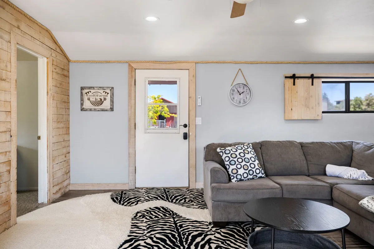 A cozy interior view with a zebra print rug, a gray sectional sofa, a wall clock, and a wooden door. The entryway area is visible, offering a glimpse of practical storage and a welcoming spot to shed outdoor gear.