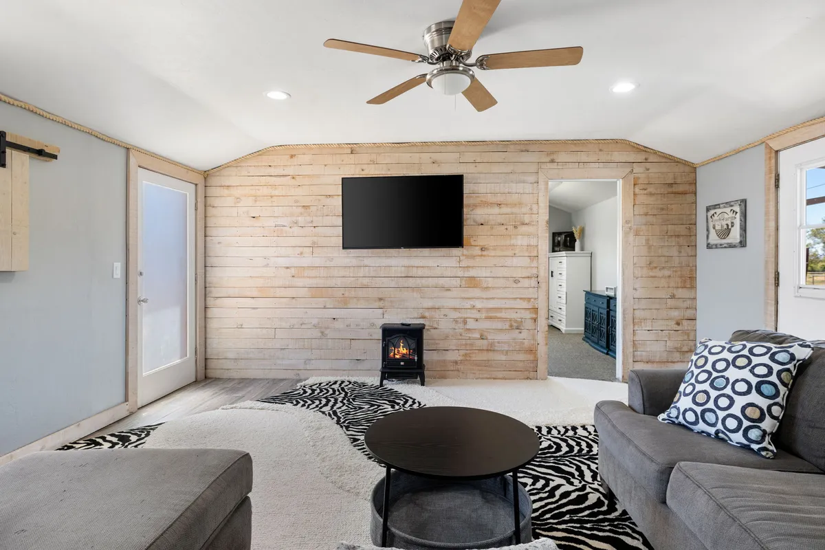 A welcoming interior lounge area showing a wooden accent wall, a fireplace, cozy armchairs and a sofa, and a television. The arrangement suggests a gathering space ideal for sipping coffee, sharing stories, or simply unwinding while looking out at the landscape beyond.