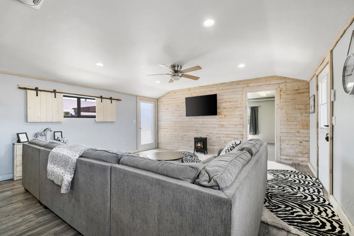 This interior image features a gray sectional sofa arranged for social gatherings, accented by a zebra print rug that adds a subtle safari vibe. The space includes wooden wall paneling and a large television, suggesting a comfortable setting for relaxing or entertaining. Bright natural light streams in, and the open layout hints at expansive windows not directly visible in the frame.